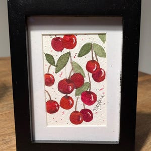 May include: A framed watercolor painting of cherries. The artwork features clusters of bright red cherries with green leaves on a white background. The painting is in a black frame with a white mat.