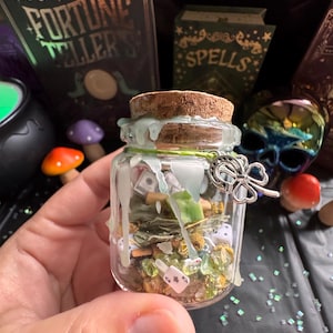 Puede incluir: Un pequeño frasco de vidrio lleno de dados, dinero y piedras verdes. El frasco está sellado con un corcho y adornado con un dije de trébol de cuatro hojas plateado. El frasco está decorado con cera azul claro.