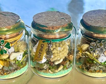 May include: Three clear glass jars with cork stoppers sealed with green wax. Each jar contains green sand, small white dice, gold-colored charms, and various trinkets. One jar features a gold tag with the word DREAM and a money bag charm.