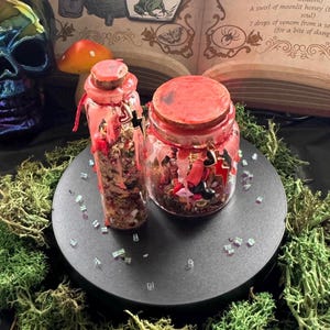 May include: Two glass containers with cork stoppers, one tall and one wide, filled with assorted small charms and beads. Both are sealed with red wax that drips down the sides. The containers feature small metal charms, including hearts and lightning bolts.