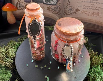 Può includere: Due barattoli di vetro trasparente con tappi di sughero, ciascuno caratterizzato da cera rosa gocciolante, uno spago arancione e un ciondolo ovale color argento. I barattoli sono riempiti con piccoli frammenti colorati su una base circolare nera.