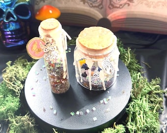 Puede incluir: Dos frascos de vidrio con tapones de corcho rellenos de pequeñas piedras y cristales de colores. Los frascos presentan gotas de cera blanca en los laterales y están adornados con dijes de metal, incluyendo un ángel y un árbol de la vida.