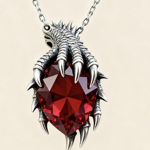 Puede incluir: Un collar con colgante plateado con una gran gema roja facetada en forma de corazón. La gema está sujeta por un diseño detallado de garra de dragón. La cadena del collar también es plateada.