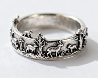 May include: A silver ring with a forest scene. The ring features detailed relief carvings of deer, a fox, trees, and mushrooms. The ring has a wide band and a slightly oxidized finish, giving it a vintage look.