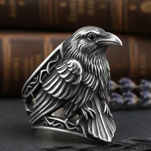 May include: A detailed silver ring depicting a raven. The bird's head and body are intricately sculpted with textured feathers and a sharp beak. The ring's band has an openwork design.