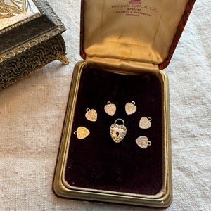 May include: A vintage jewelry box with a red velvet interior holds several small silver heart-shaped charms and a heart-shaped padlock. The box is open, revealing the charms against the dark background. The box is trimmed with gold.