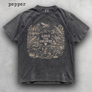 May include: A charcoal grey short-sleeve t-shirt featuring a vintage-style graphic of various insects and plants surrounding the text LOVE THE UNLOVED with a small broken heart icon. The shirt has a faded, washed-out appearance and a crew neckline.