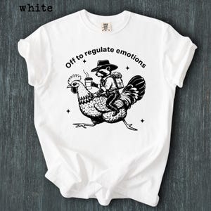 May include: White t-shirt featuring a black and white illustration of a raccoon riding a chicken, holding a coffee cup, and wearing a hat and backpack. The text "Off to regulate emotions" is above the graphic.