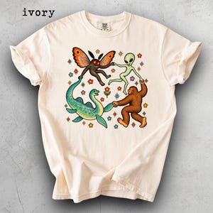 May include: An ivory short-sleeve t-shirt featuring a graphic print of a moth, an alien, a Loch Ness monster, and a Bigfoot holding hands in a circle. The design includes colourful small flowers and star shapes surrounding the figures. The word ivory is printed in the top left corner.