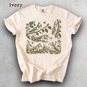 May include: An ivory t-shirt featuring a dark green graphic print of multiple praying mantises in various poses among leaves and branches. The word ivory is printed in the top left corner. The shirt has a crew neckline and short sleeves.