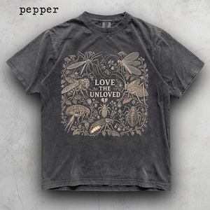 May include: A pepper-coloured short-sleeve t-shirt featuring a graphic print of various insects and botanical elements. The text "LOVE THE UNLOVED" is centred in the design, accompanied by a small broken heart icon above the central insect illustration.