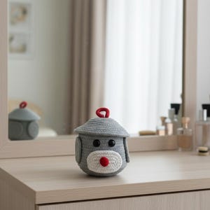 May include: A gray crocheted dog-shaped container with a red loop on top. The container has a white muzzle with a red nose and black eyes. The container is sitting on a light-colored wooden surface.