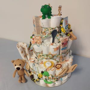 Könnte beinhalten: Ein dreistöckiger Windelkuchen, verziert mit Babyartikeln. Der Kuchen ist mit einem grünen Frosch, einer Giraffe, einem Teddybären, einem Schnuller und verschiedenen Babyprodukten geschmückt. Der Kuchen ist mit gemusterten Bändern umwickelt und mit Aufklebern zum Thema Tiere verziert.