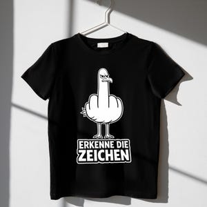 Puede incluir: Camiseta negra con un dibujo de una gaviota blanca haciendo una peineta. El texto "ERKENNE DIE ZEICHEN" se muestra en un rectángulo blanco debajo. La camiseta está colgada en una percha blanca.