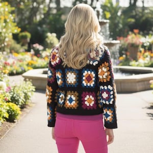 Può includere: Una giacca nera all'uncinetto con motivi quadrati colorati, indossata con pantaloni rosa. La giacca presenta quadrati nelle tonalità di bianco, arancione, blu e viola. Lo sfondo mostra un giardino con una fontana.
