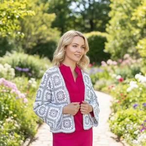 Op de afbeelding: Een witte en grijze gehaakte jas met een bloemenpatroon. De jas is open en onthult een felroze top. De achtergrond is een tuin met een pad en kleurrijke bloemen.