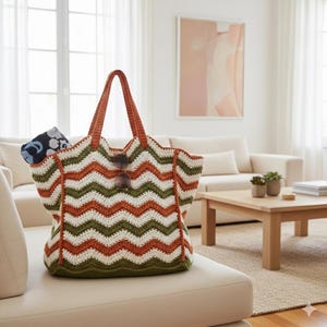 May include: A large, handmade crochet tote bag with a chevron pattern in white, olive green, and orange. The bag has brown handles and a pair of sunglasses hanging from the front. A patterned scarf is visible inside the bag. The bag is sitting on a white couch.
