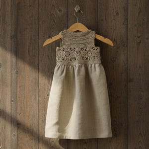 May include: A beige sleeveless dress with a crocheted bodice and a flowing skirt, hanging on a wooden hanger. The bodice features a floral crochet pattern. The dress is made of a soft, natural-looking fabric, ideal for a summer day.