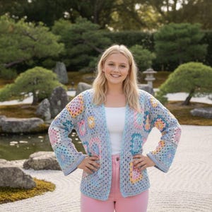 May include: A light blue, open-front crochet cardigan with colorful floral squares. The cardigan features pastel pink, yellow, and blue squares. The sleeves are long and flared. The person is wearing a white top and pink pants.