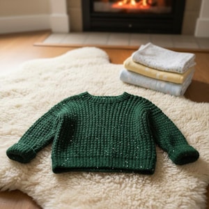 May include: A dark green knit sweater with white flecks, lying on a cream-colored, textured rug. The sweater has a ribbed texture and folded cuffs. In the background, a stack of folded blankets and a fireplace with a lit fire are visible.