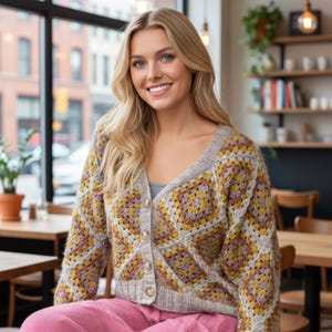 Puede incluir: Una mujer con un cárdigan abotonado. El cárdigan presenta un diseño de cuadrados de la abuela en tonos amarillos, marrones y grises. La mujer también lleva una camisa gris y pantalones rosas. El fondo muestra un café.