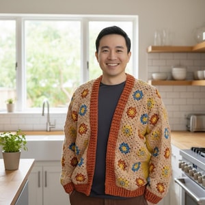 Könnte beinhalten: Beige Häkel-Cardigan mit einem Blumenmuster in Gelb, Blau und Orange. Der Cardigan hat eine rostfarbene Bordüre. Die Person trägt ein graues Hemd. Der Hintergrund zeigt eine Küchenumgebung.