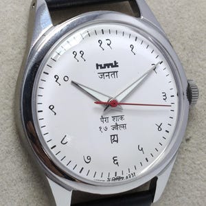 May include: A silver-toned wristwatch with a white face and black leather band. The watch face features black numerals, the brand name "humt जनता", and red second hand. The watch has a classic, minimalist design.