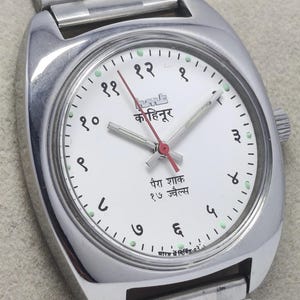 May include: A silver-toned wristwatch with a white face and a matching silver band. The watch face features black numerals, green dots, and red hands. The watch face has text in a foreign language.