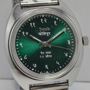 May include: A silver-toned wristwatch with a green dial and Arabic numerals. The watch face includes text in a foreign language. The watch has a stainless steel band.