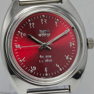 May include: A silver-toned wristwatch with a red dial featuring Hindi numerals and text. The watch has a mesh band and a crown on the right side. The dial includes the text "कोहिनूर" and "पैरा शाक १७ ज्वैल्स".