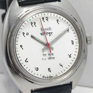 May include: A silver-toned wristwatch with a white face and black leather band. The watch face features black numerals, green dots, and text in a foreign language. The watch hands are silver and red.