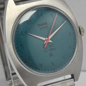May include: A vintage wristwatch with a teal face and silver-tone metal band. The watch face features Arabic numerals and text in a foreign language. The hands are silver with a red second hand.