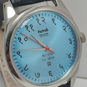 May include: A wristwatch with a light blue face, silver casing, and a dark blue leather band. The watch face features Hindi numerals and text, with red and silver hands. The watch is marked "hmt जनता" and "पैरा शाक १७ ज्वैल्स".