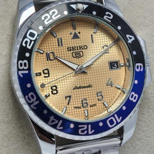 May include: A stainless steel Seiko 5 automatic watch with a gold-colored dial and a blue and black bezel. The watch face displays the time with luminous hands and markers, and the date is visible at the 3 o'clock position.