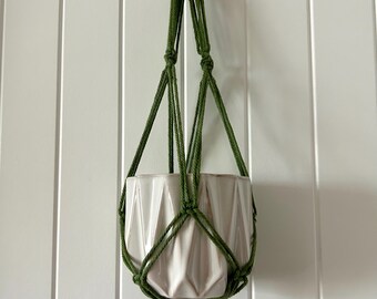 May include: A green macrame plant hanger with a white geometric ceramic pot. The hanger is suspended from a wooden ring. The pot has a textured, angular design, and the macrame has a tassel at the bottom.