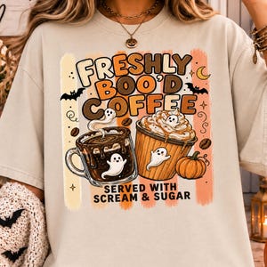 May include: A beige short-sleeve t-shirt featuring a graphic print with the text "FRESHLY BOO'D COFFEE SERVED WITH SCREAM & SUGAR". The design includes two coffee cups, ghosts, bats, coffee beans, and a pumpkin in shades of brown and orange.
