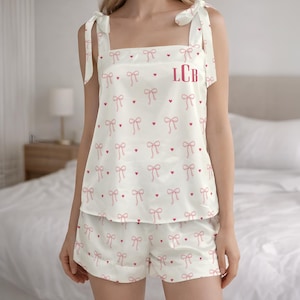 Puede incluir: Conjunto de pijama color crema con un estampado de lazos y corazones rosas. La parte superior tiene tirantes anudados y las iniciales "LCB" en rosa. Los pantalones cortos coinciden con el diseño de la parte superior. El conjunto es de material sedoso.