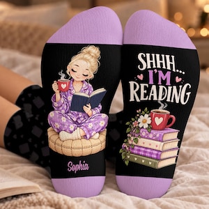 Puede incluir: Un par de calcetines negros con puños color lavanda. Uno muestra un personaje rubio en pijama floral morado leyendo un libro con el nombre Sophia debajo. El otro muestra una pila de libros y una taza roja con el texto SHHH... I'M READING.