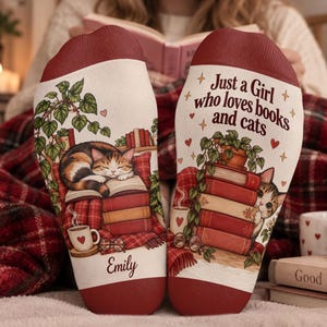 Può includere: Un paio di calzini color crema con polsini rosso scuro. Il calzino sinistro mostra un gatto calico che dorme sui libri, una pianta in vaso e il nome Emily. Il destro mostra un gatto dietro dei libri, una pianta e il testo Just a Girl who loves books and cats.