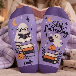 May include: A pair of light purple socks featuring a ghost wearing glasses and reading a book while sitting on a stack of books, with a pumpkin, stars, and a crescent moon. The right sock includes the text "Shhh I'm reading" and the left says "Emily".