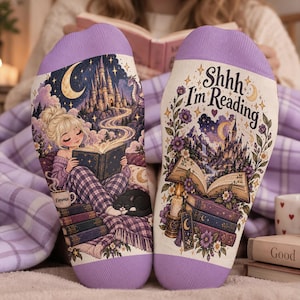 Pode incluir: Um par de meias com punhos lavanda com uma estampa gráfica de uma pessoa lendo um livro com um gato, um castelo e uma lua crescente. A outra meia exibe o texto Shhh I'm Reading acima de um livro aberto e uma pilha de livros.