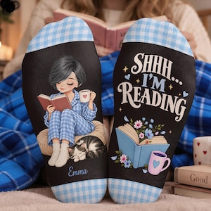 Puede incluir: Un par de calcetines negros con un patrón de cuadros vichy azul claro en la parte superior e inferior. Un calcetín presenta una ilustración de una persona leyendo un libro con un gato y el nombre Emma. El otro calcetín muestra el texto SHHH... I'M READING con un libro y una taza.