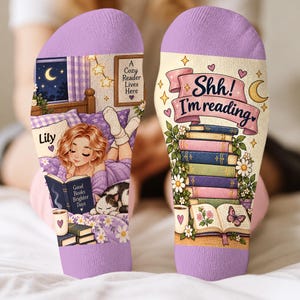 May include: A pair of lavender-cuffed socks featuring a printed illustration of a person reading in bed with a cat, the text 'Lily', 'Good Books Brighter Days', 'A Cozy Reader Lives Here', and 'Shh! I'm reading' above a stack of colourful books.