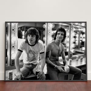 Puede incluir: Una fotografía enmarcada en blanco y negro que muestra a dos personas sentadas en un camerino con luces de espejo. Una persona lleva una camiseta de manga corta y vaqueros, mientras que la otra no lleva camisa y viste vaqueros. Ambas sostienen bebidas.