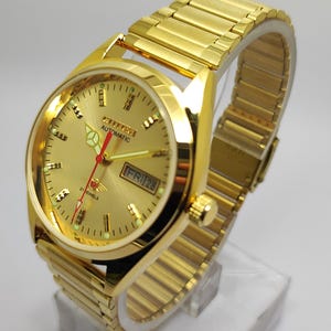 May include: Gold-coloured Citizen automatic wristwatch with a matching gold strap. The watch face shows the day and date, with a gold dial, black markers, and red hands. The watch is displayed on a clear stand.