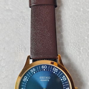 May include: A gold-coloured Seiko Quartz watch with a blue dial and brown leather strap. The watch face displays the time with white numerals and markers. The strap is a dark brown colour.