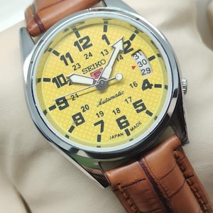 Peut inclure: Une montre-bracelet automatique Seiko avec un cadran jaune et un bracelet en cuir marron. La montre a un boîtier argenté et présente des chiffres et des aiguilles noirs. Le cadran affiche le jour du mois et le texte "Automatic" et "Japan Made".