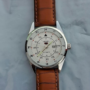 May include: A silver-toned Seiko automatic wristwatch with a white face and brown leather band. The watch face displays the day and date, with black numerals and hands. The watch has a classic, vintage design.