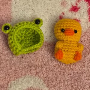 May include: A handmade crochet set featuring a lime green frog hat with black eyes and a yellow chick with orange feet and beak. The toys are made of yarn and are on a pink and white textured surface.