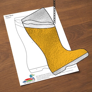May include: A stained glass pattern for a beer boot, with a chain for hanging. The boot is filled with golden yellow glass, with a silver-coloured top and sole. The pattern is on a white sheet of paper with the text "Beer Boot Pattern".
